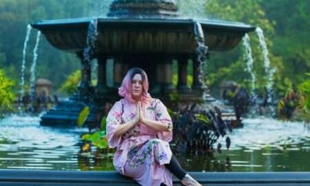 Person in pink traditional clothing sitting cross-legged with hands in prayer position in front of a fountain with water flowing.