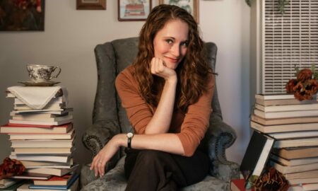 Young woman with curly hair sitting in a gray armchair surrounded by stacks of books, smiling and resting her chin on her hand.