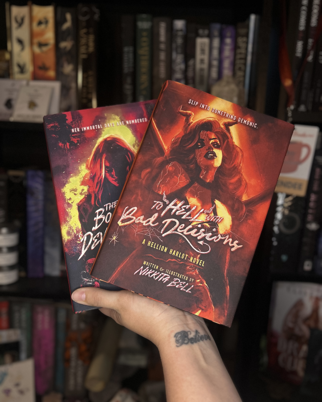 Two illustrated book covers held in a person's hand, featuring fiery and dark themes with female characters.