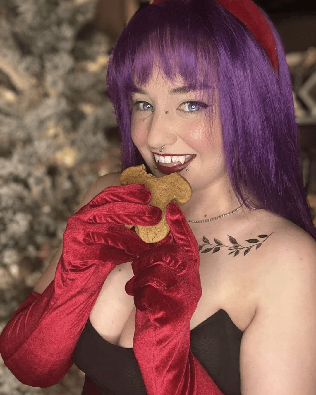 Woman with purple hair and red gloves holding a cookie, smiling, in front of a Christmas tree.
