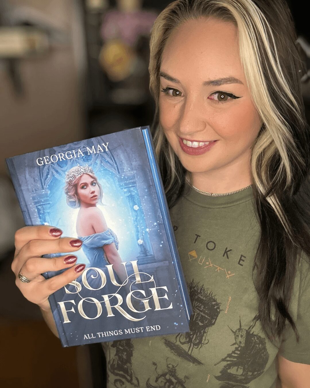 Young woman with long blonde hair smiling, holding a book titled 'Soul Forge' by Georgia May, in a cozy setting.