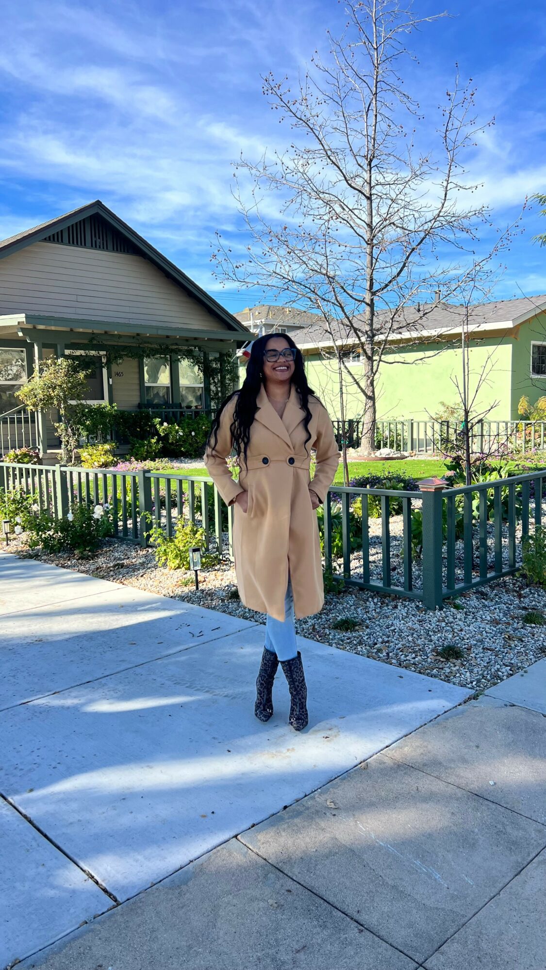Woman standing outdoors in front of a green building and leafless tree, wearing a beige coat and black boots.