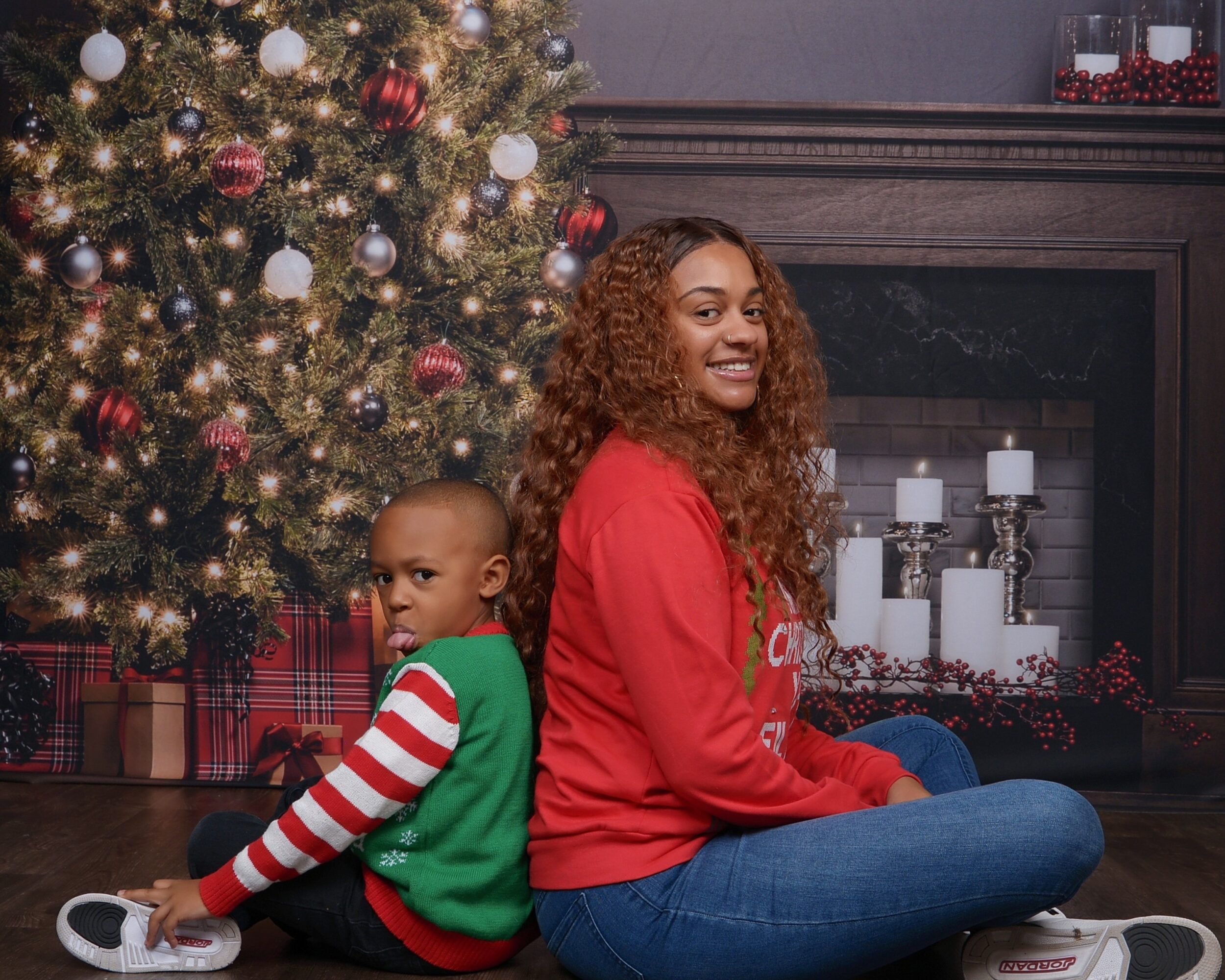 Two children sitting on the floor in front of a decorated Christmas tree and fireplace, smiling.