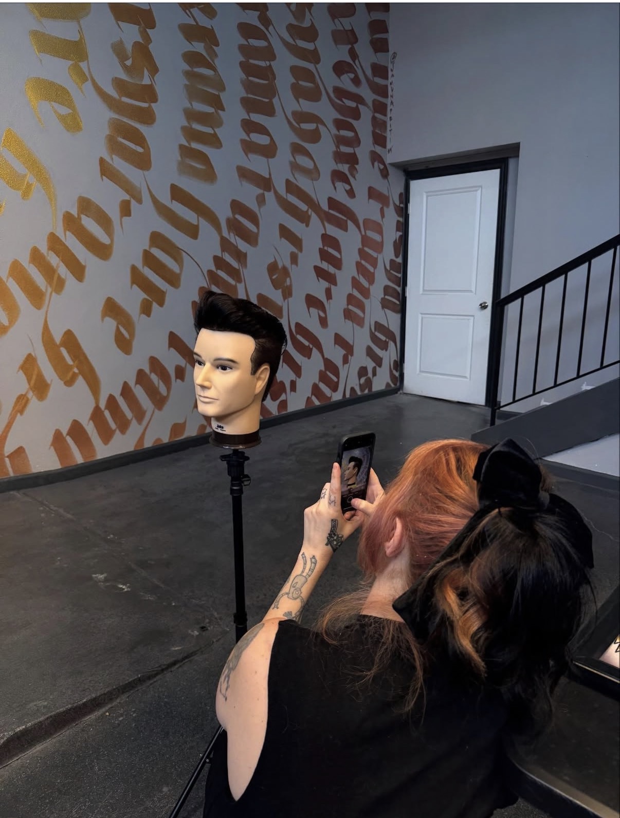 Person with pink hair taking a photo of a mannequin head on a stand in a room with wall text.