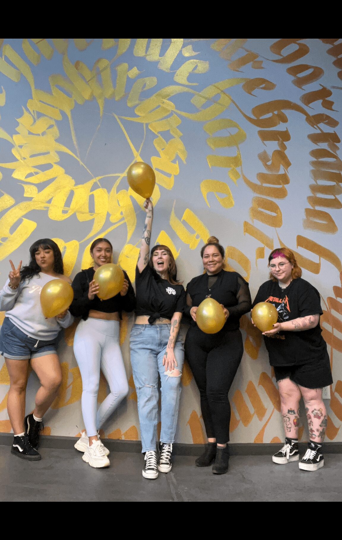 Five women standing in front of a wall with large gold and blue text, holding gold balloons, smiling and posing.