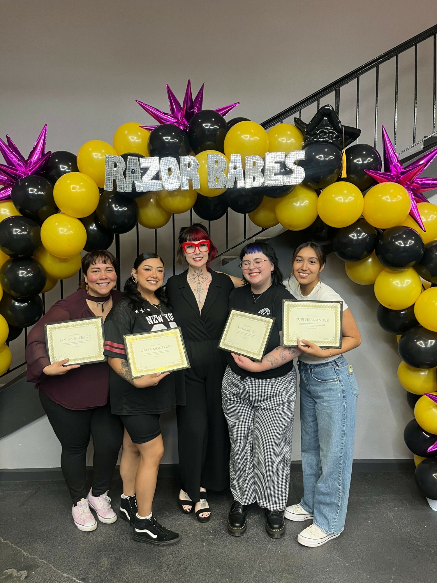 Five women standing in front of a balloon arch with a sign that reads 'RAZOR BABES'. They are holding certificates and smiling.