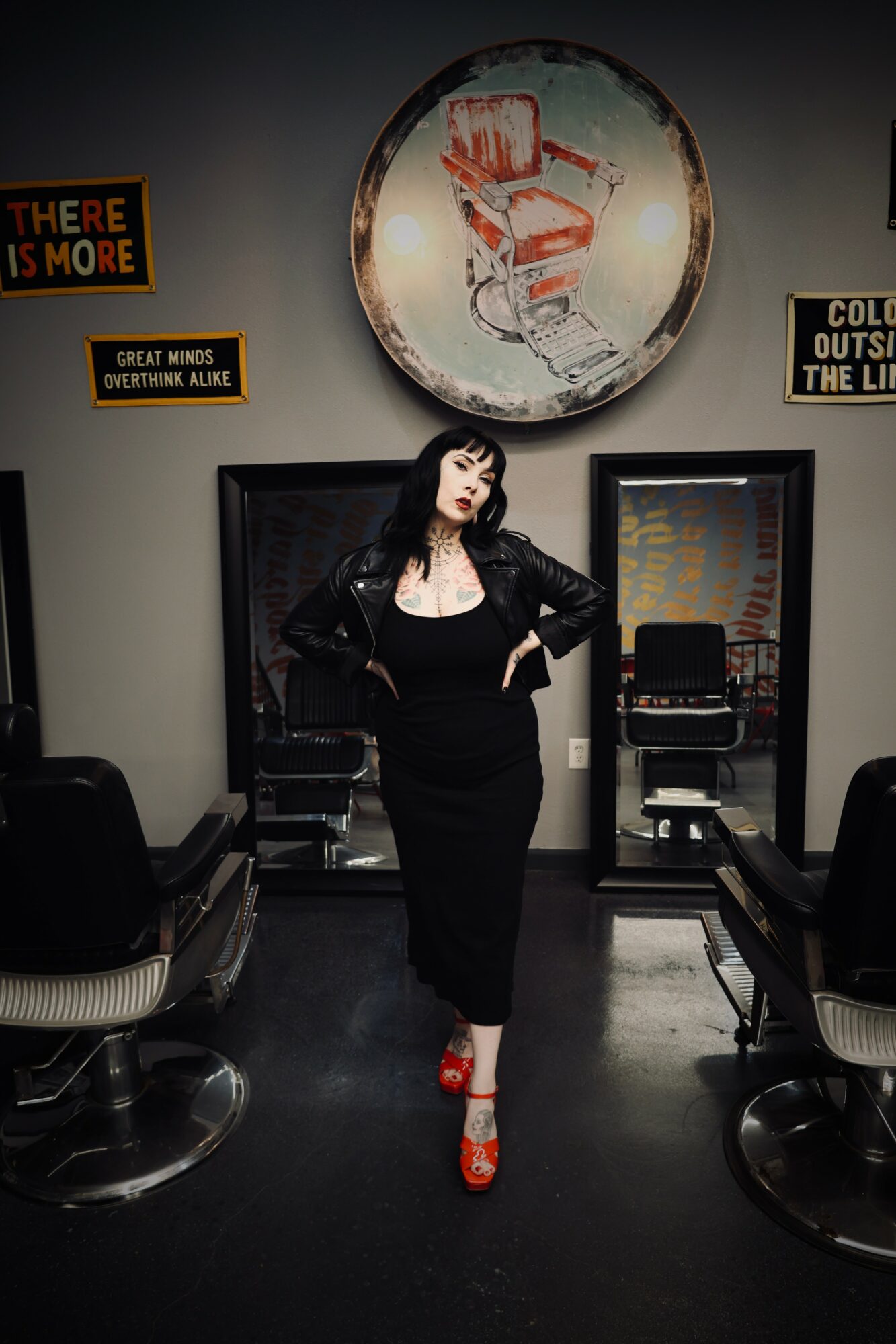 Woman in black dress and red shoes standing in salon with chairs and mirrors, large circular artwork above her head.