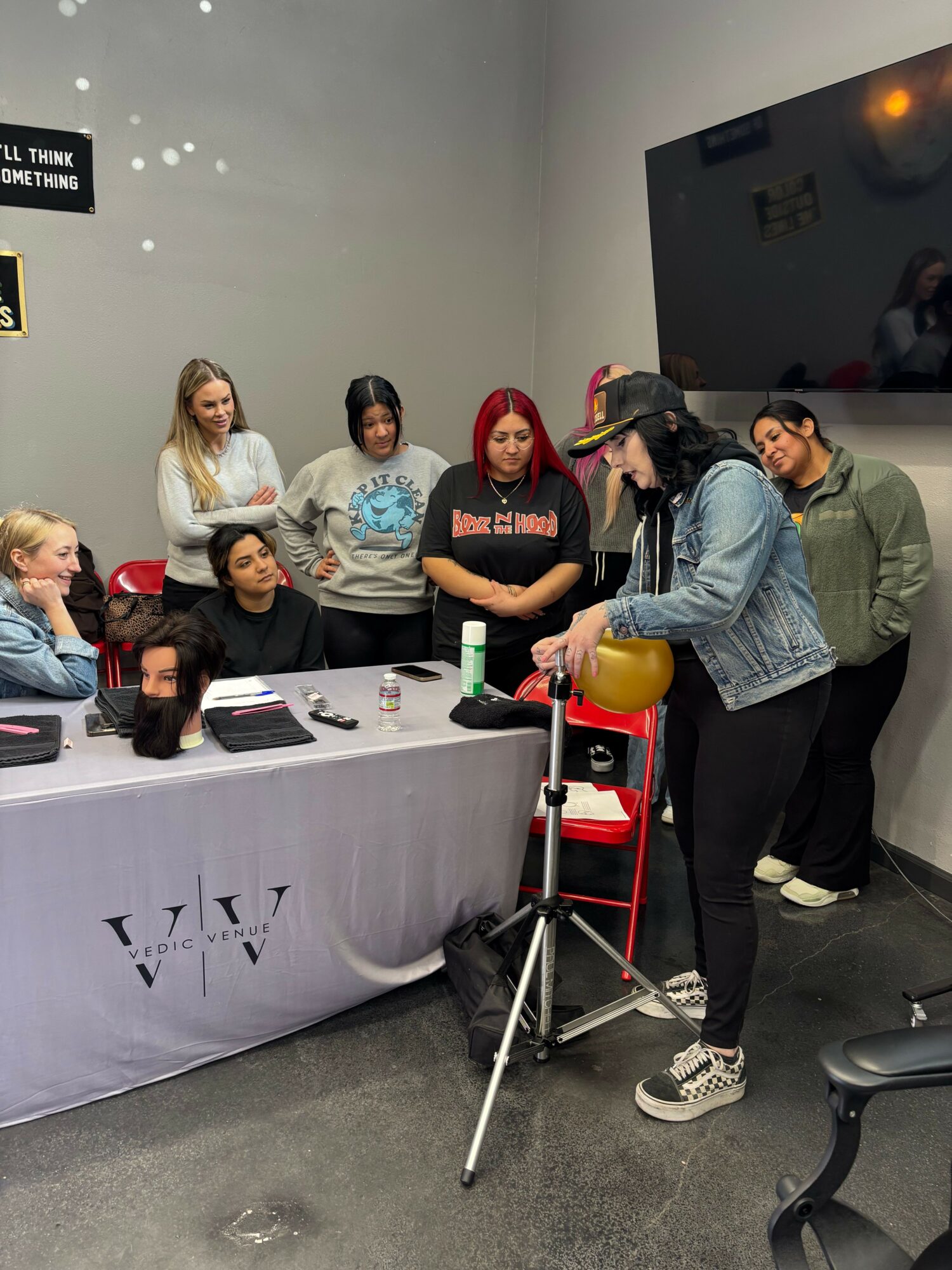 Group of people watching a woman demonstrate using a yellow device on a tripod in a room with a large screen.
