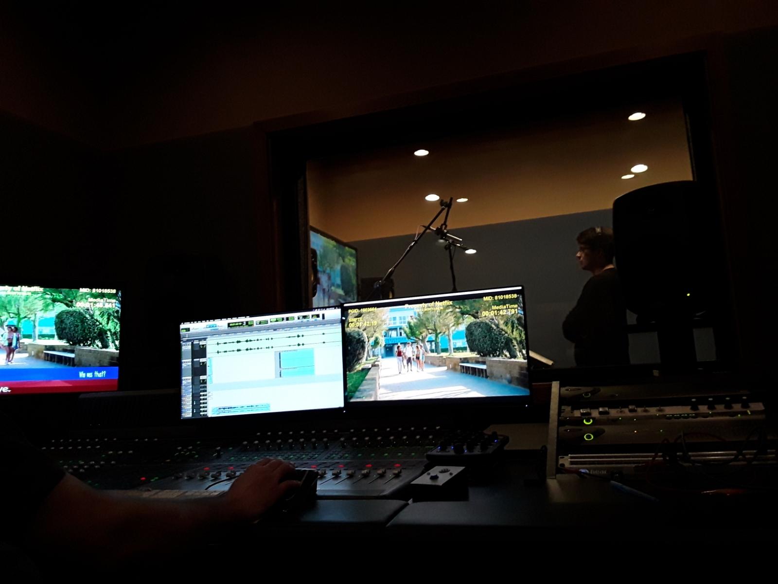 Recording studio with two computer monitors displaying outdoor scenes, a person working, and audio equipment in a dimly lit room.