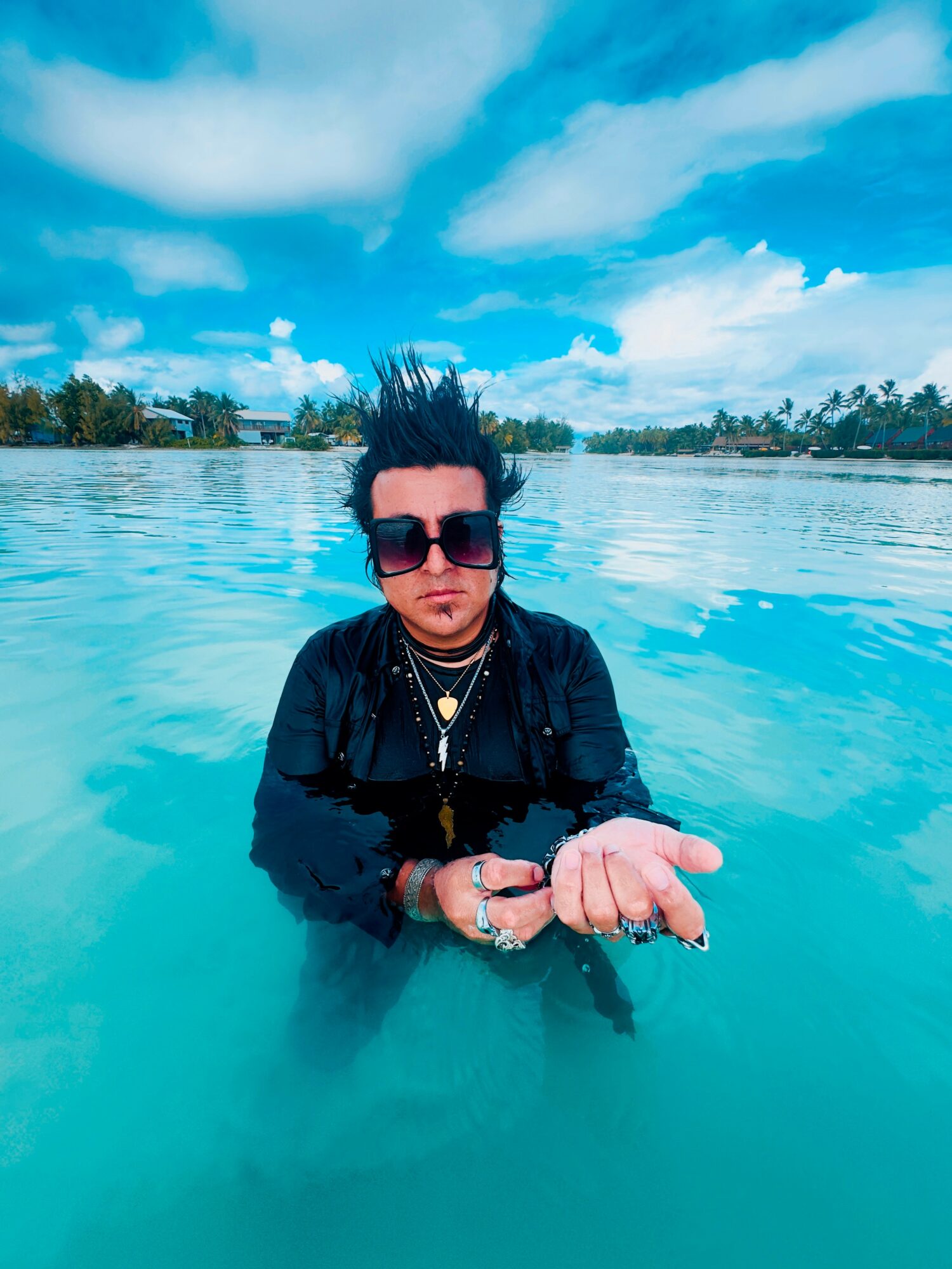 Person with dark hair and sunglasses standing in water, holding their hands together, with a cloudy sky and trees in background.