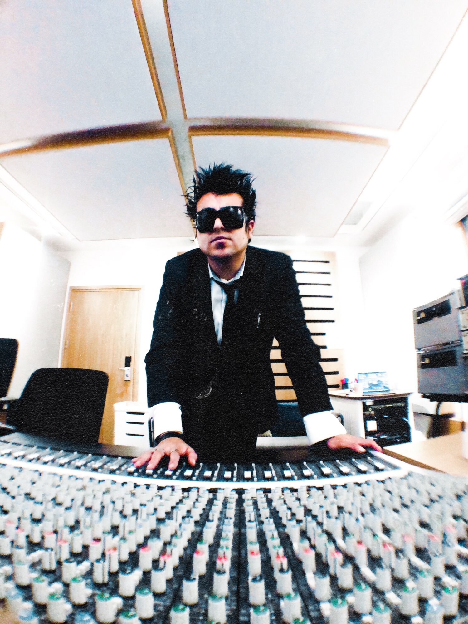 Person in sunglasses and suit leaning over mixing console in recording studio.