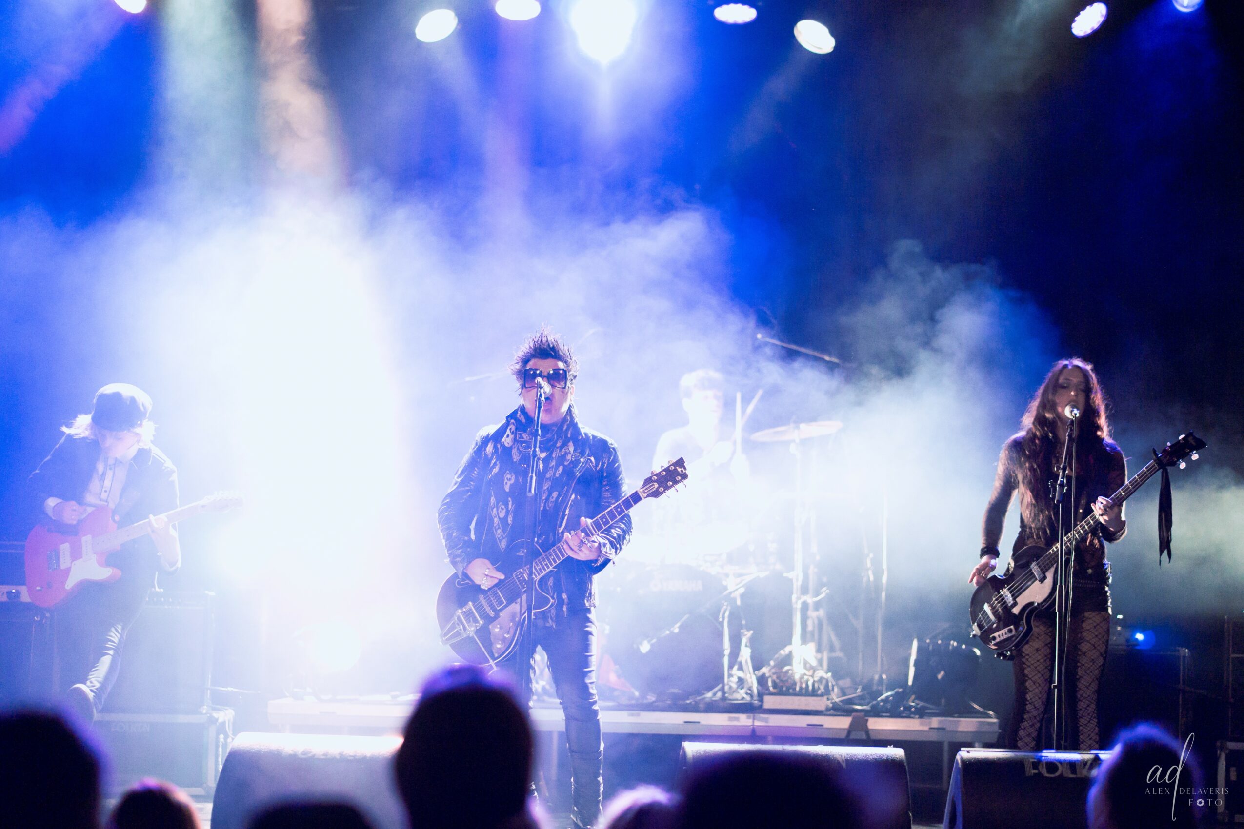 Band performing on stage with bright lights and fog, three musicians playing guitars and drums, audience in foreground.