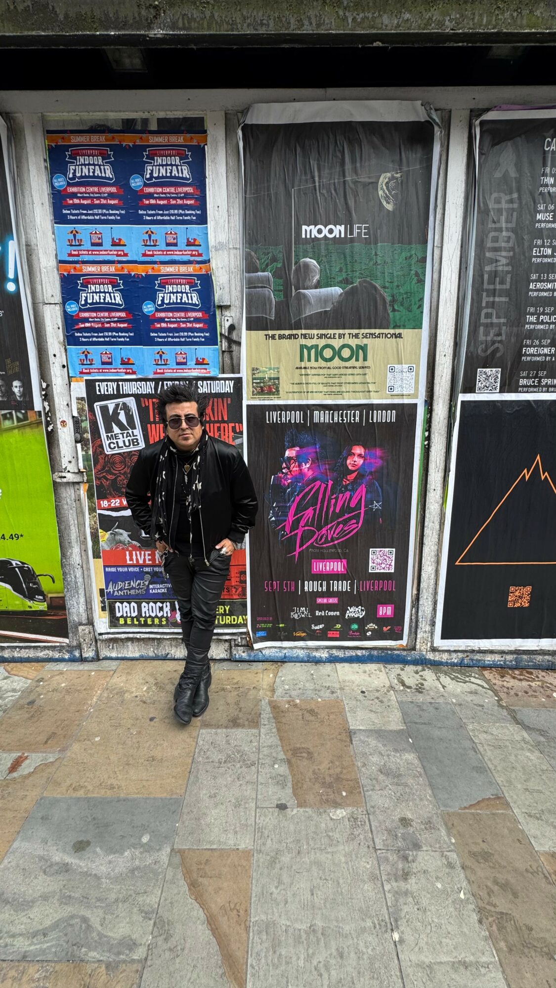Person in black jacket and sunglasses standing in front of a wall with posters and advertisements.