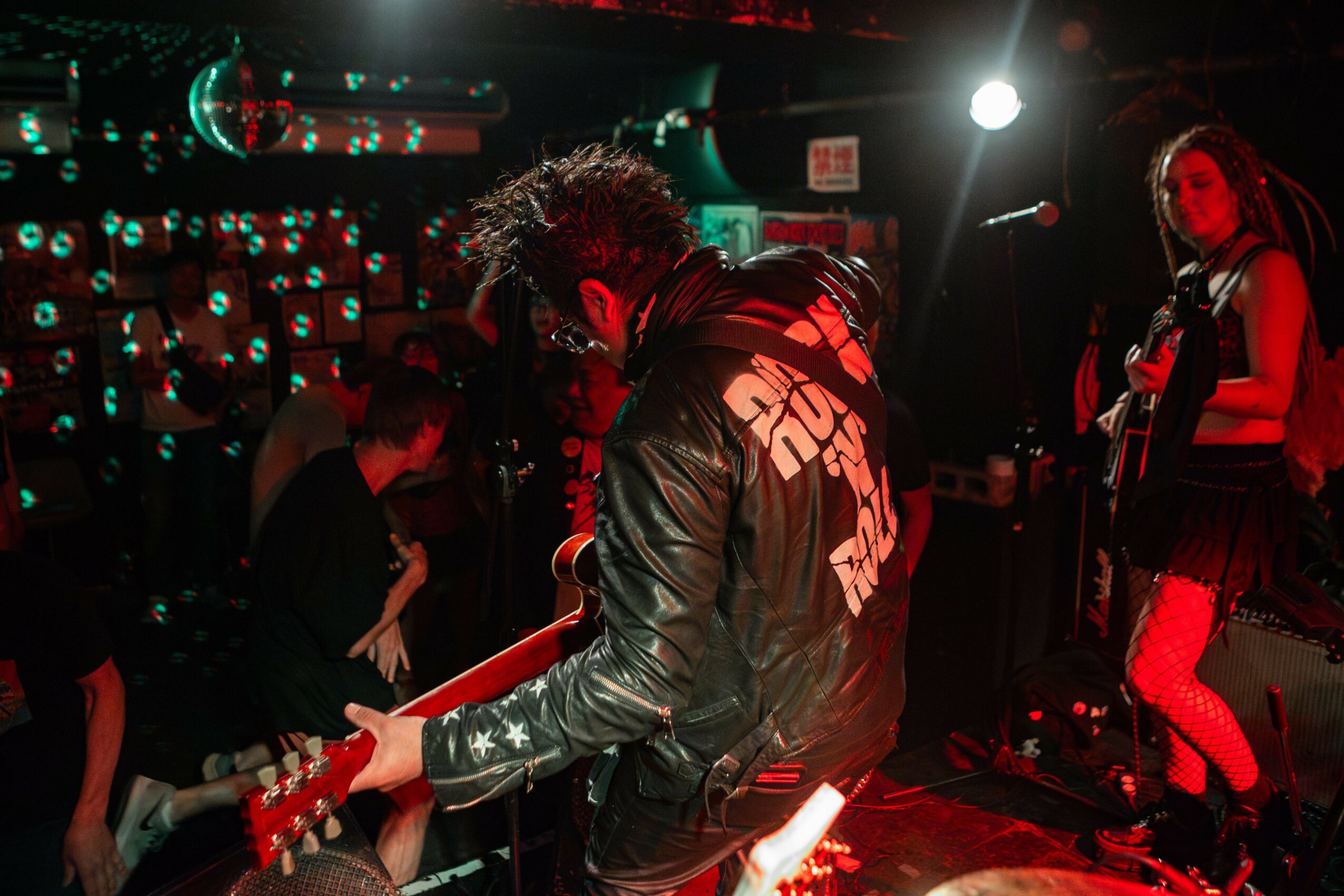 Musician playing guitar on stage with a woman singing in the background, dimly lit with colorful stage lights.