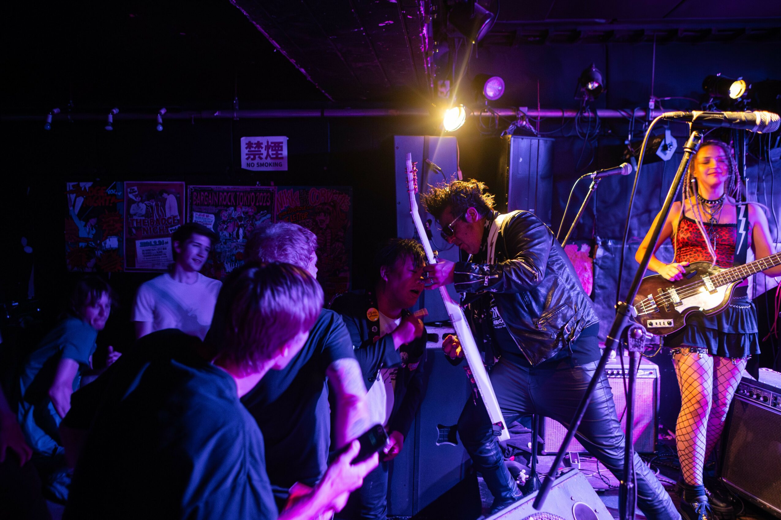 Musician with curly hair and leather jacket performs on stage with a woman playing guitar, audience watching attentively.