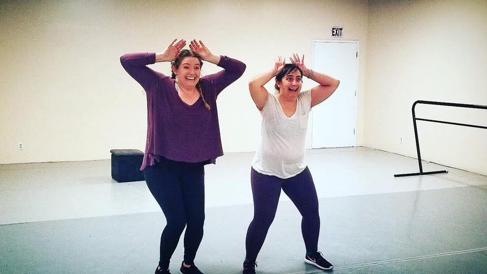 Two women stand in a dance studio, smiling and holding their hands above their heads in a playful pose.