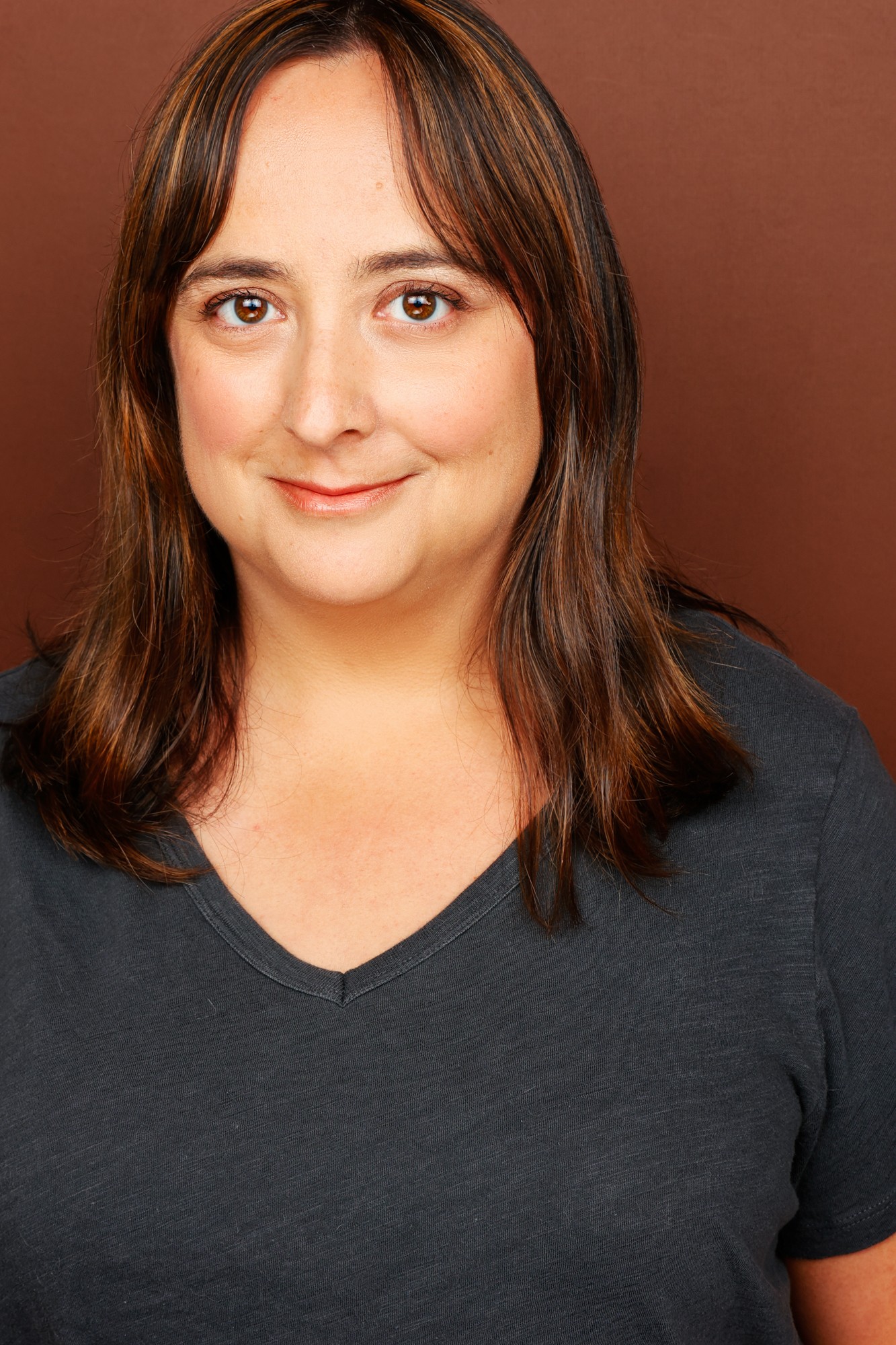 Woman with shoulder-length brown hair, smiling, wearing a dark top, against a brown background.