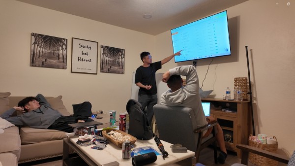 Two people in a living room, one presenting data on a wall-mounted screen, another sitting at a table, a third on a sofa.