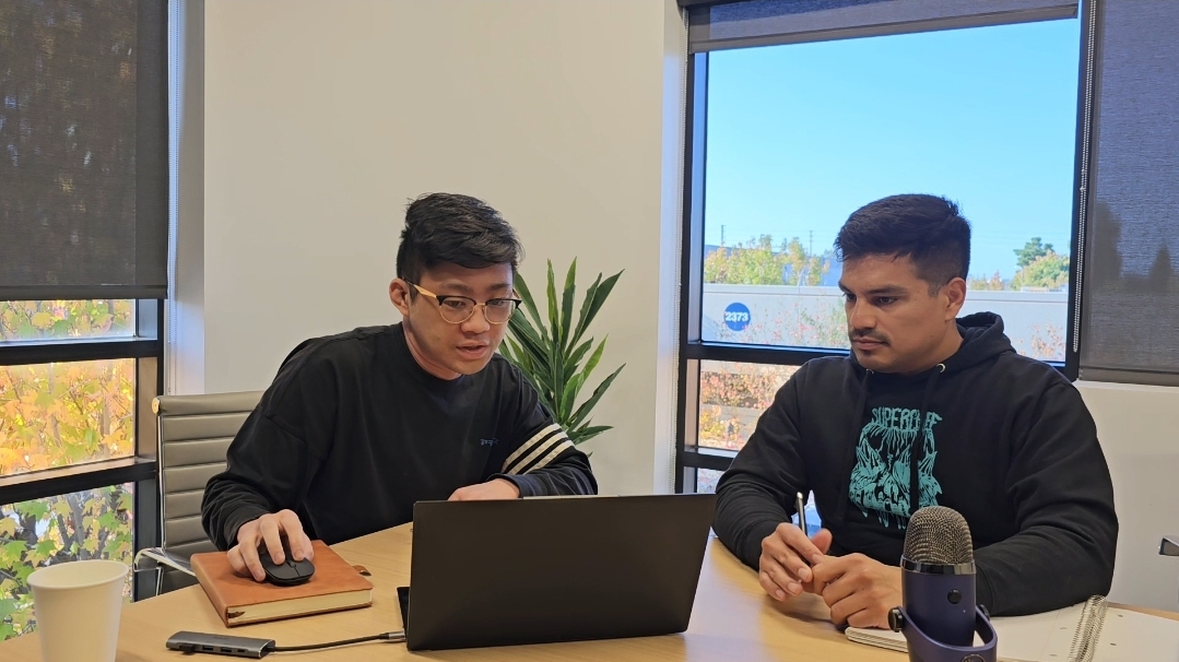Two men sit at a table with a laptop, a plant, and a microphone, in a room with large windows showing a blue sky.