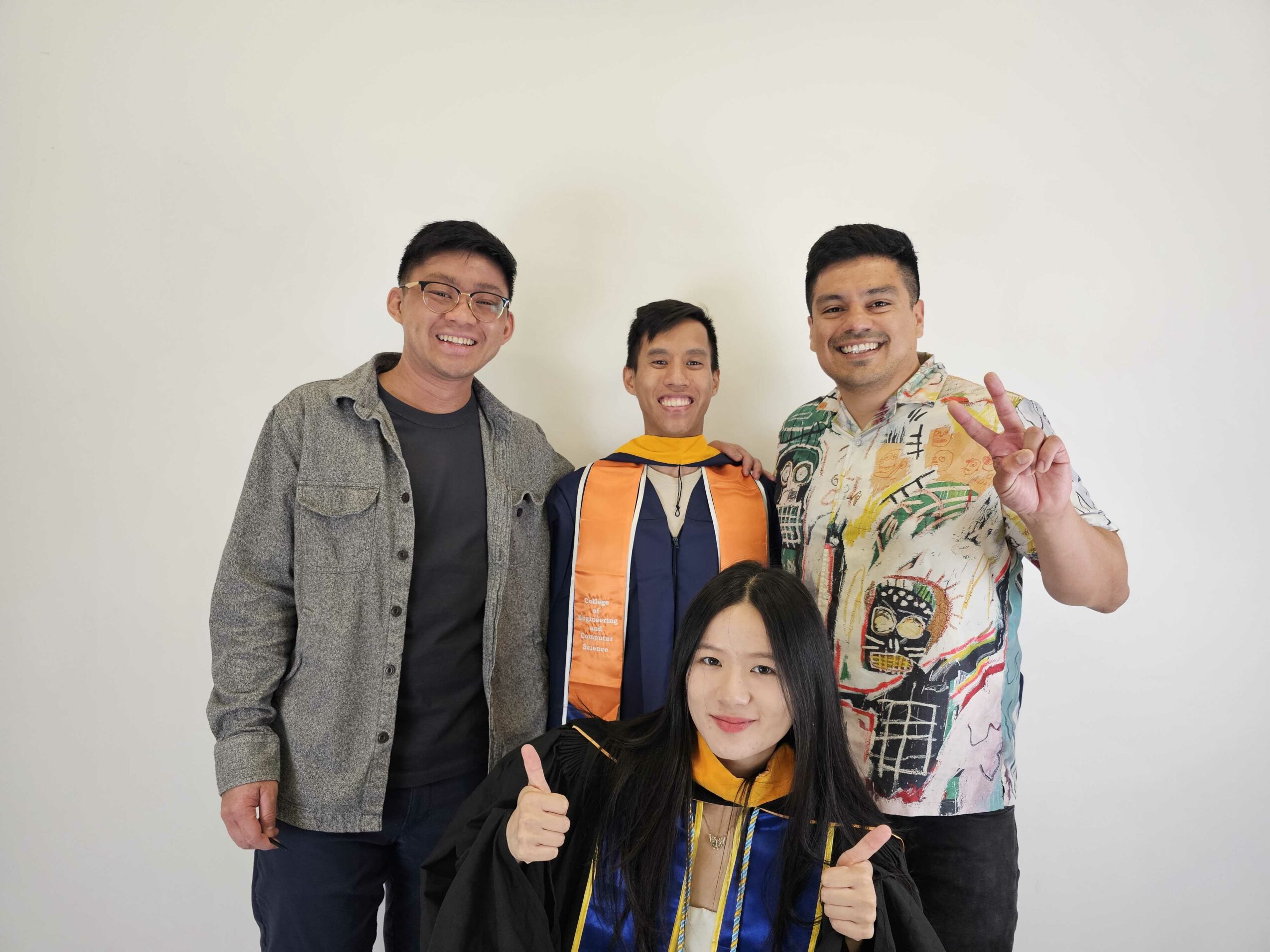 Four people standing together, smiling, two giving thumbs up, one making a peace sign, against a plain white background.