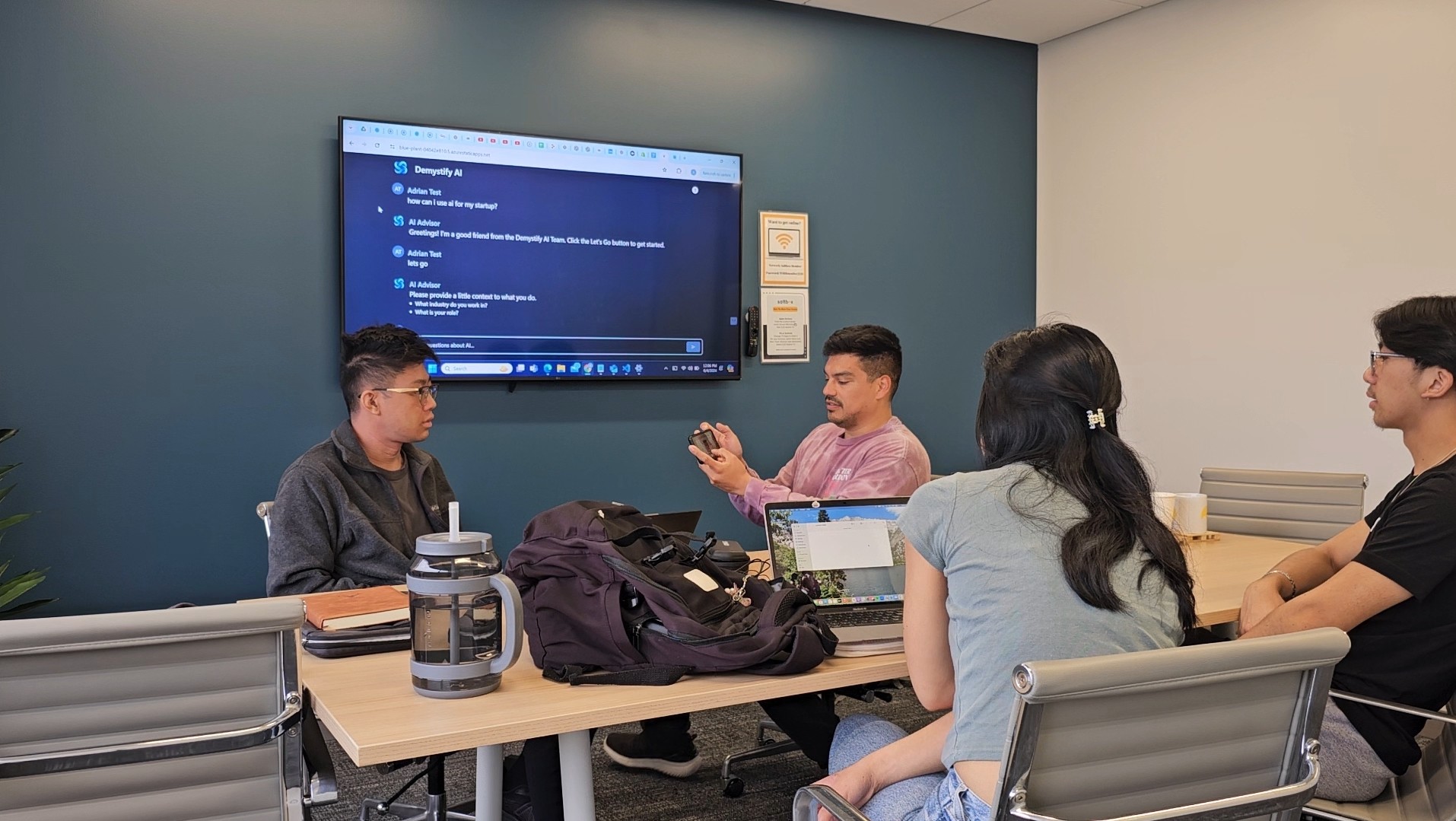 Group of five people sitting around a table in a meeting room, with a large screen on the wall displaying a presentation.