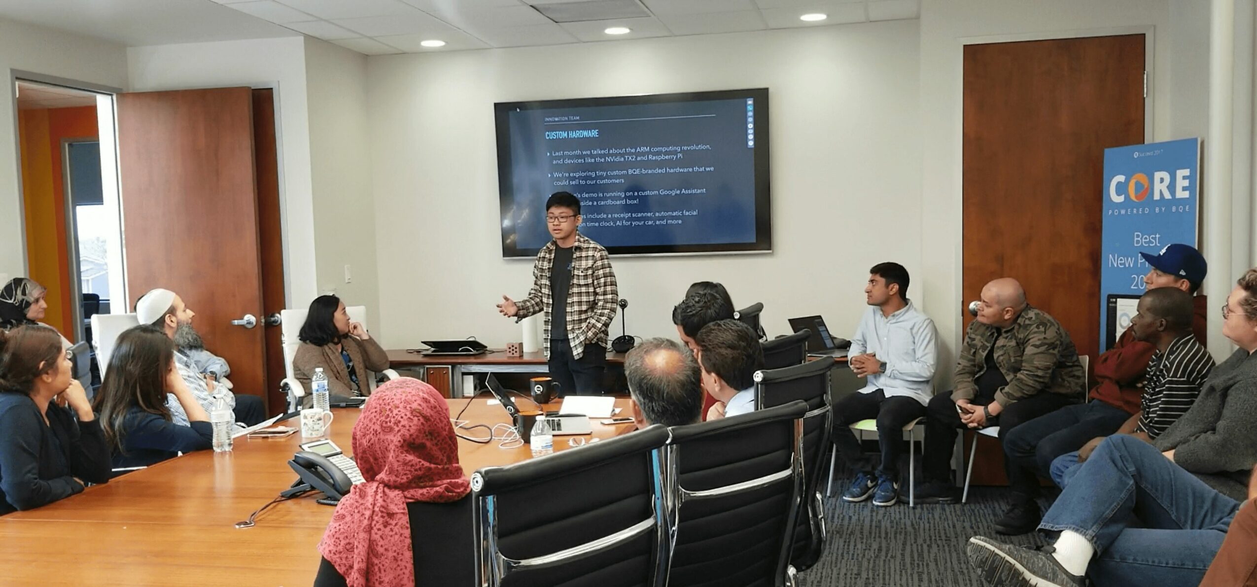 Person standing and presenting to seated group in conference room with large screen behind.