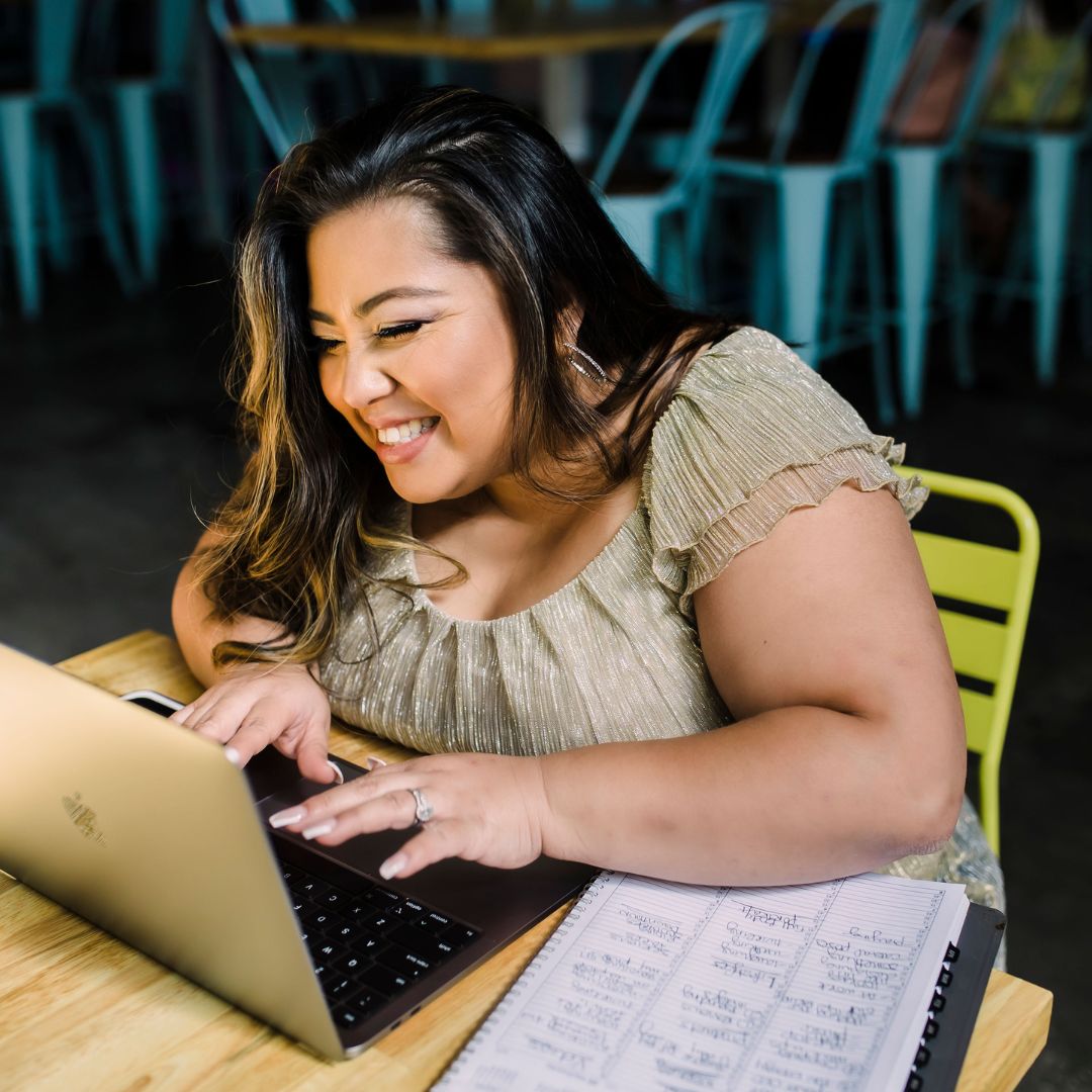 Woman with long dark hair smiling and typing on a laptop at a wooden table.