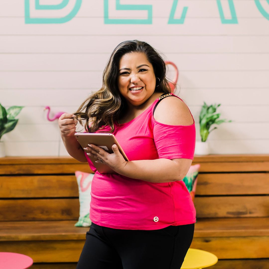Smiling woman with long wavy hair in pink top holding a tablet, standing in front of a decorated wall.