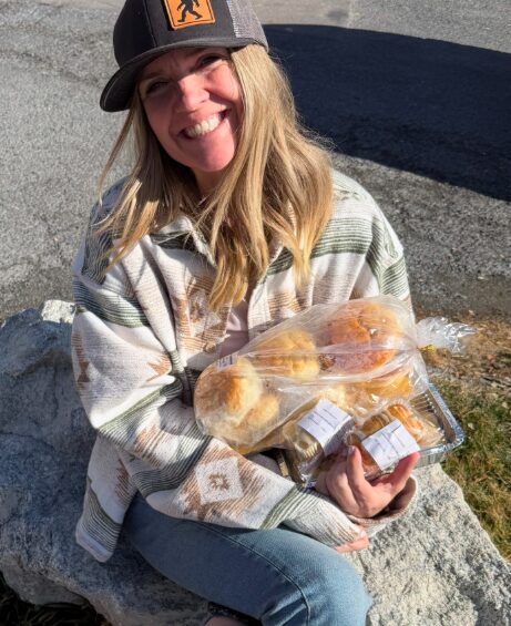 Person with long hair wearing a cap and striped sweater holding a tray of bread outdoors.