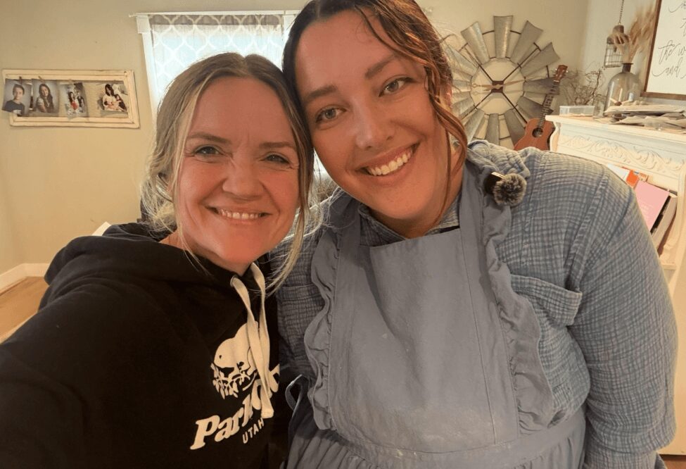 Two women smiling indoors, one wearing a black hoodie and the other in a light blue apron, in a cozy room.