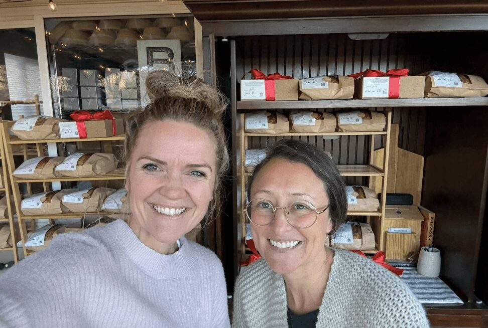 Two women smiling in front of shelves with bread and bakery items.