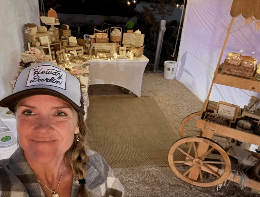 Woman taking a selfie at an outdoor market stall with baskets and wooden carts, evening setting with lights and display tables.