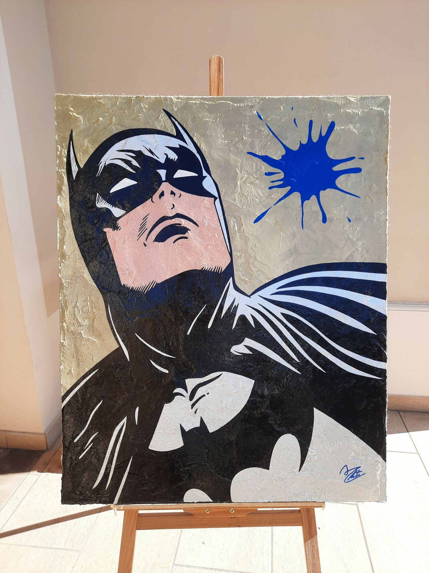 Painting of Batman with a blue splatter above his head, on a textured background, on an easel.