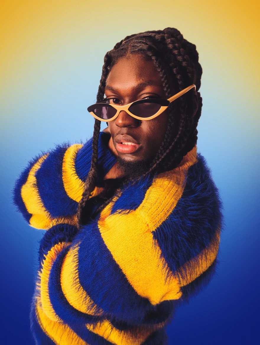 Woman with braided hair wearing yellow and blue striped sweater and sunglasses, posing with hand near face.