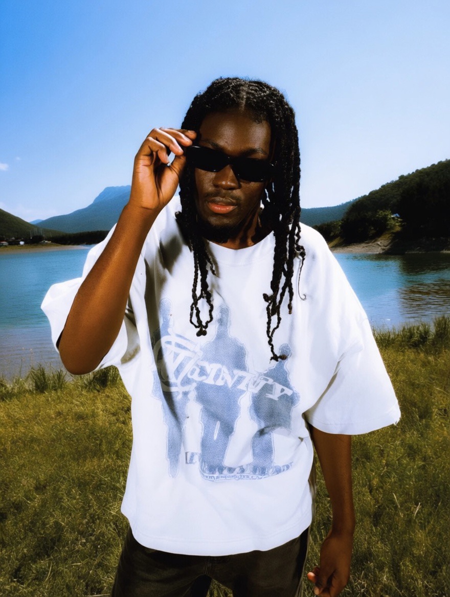 Person with long dreadlocks wearing sunglasses and a white graphic t-shirt outdoors near water and green hills.
