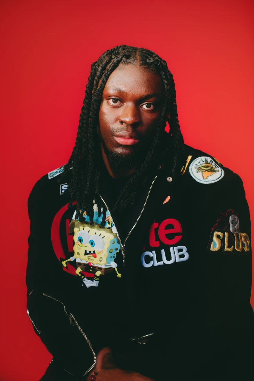 Person with braided hair wearing a black jacket with patches and colorful designs, against a red background.