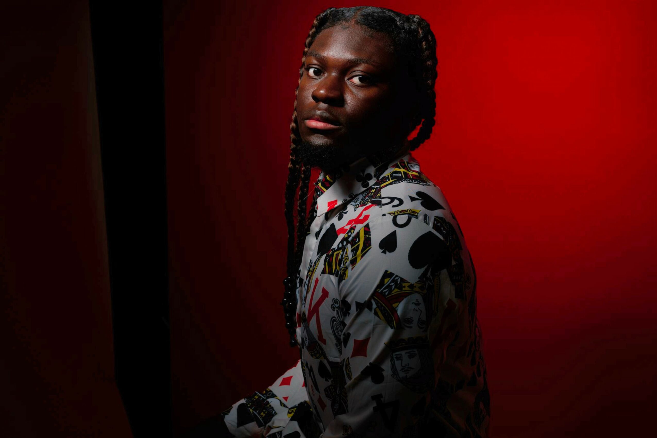 Person with braided hair wearing a patterned shirt, standing against a red background, looking at the camera.