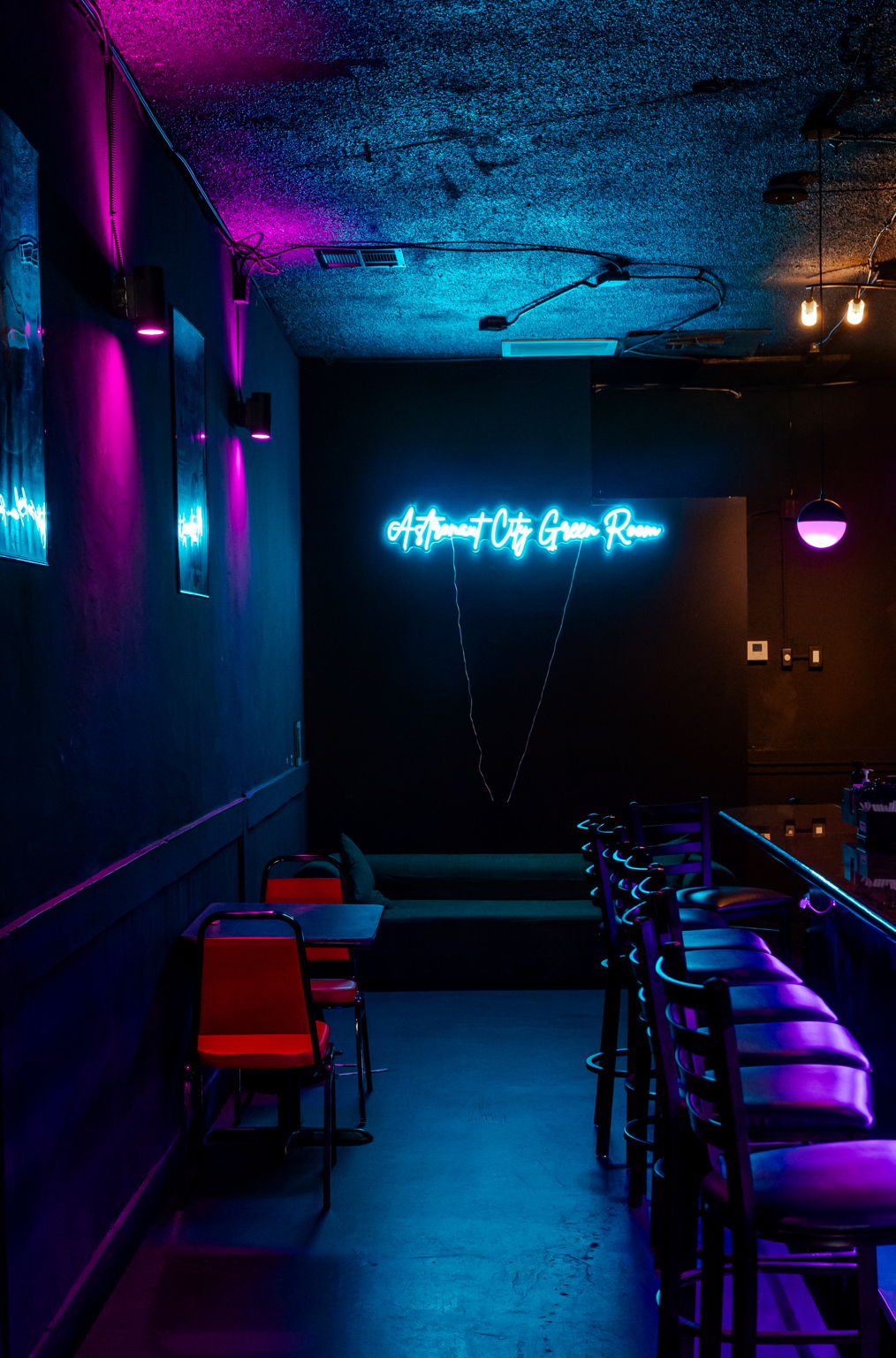 Neon sign on dark wall, purple and blue lighting, chairs and tables in a dimly lit room.