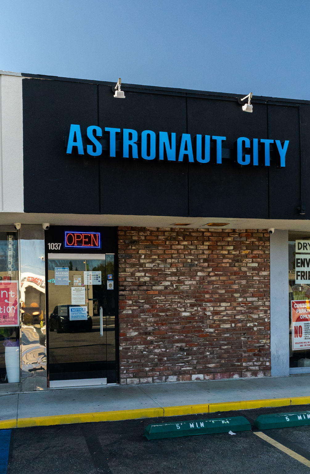 Storefront with blue sign reading 'ASTRONAUT CITY' above a brick wall and glass door with an open sign.