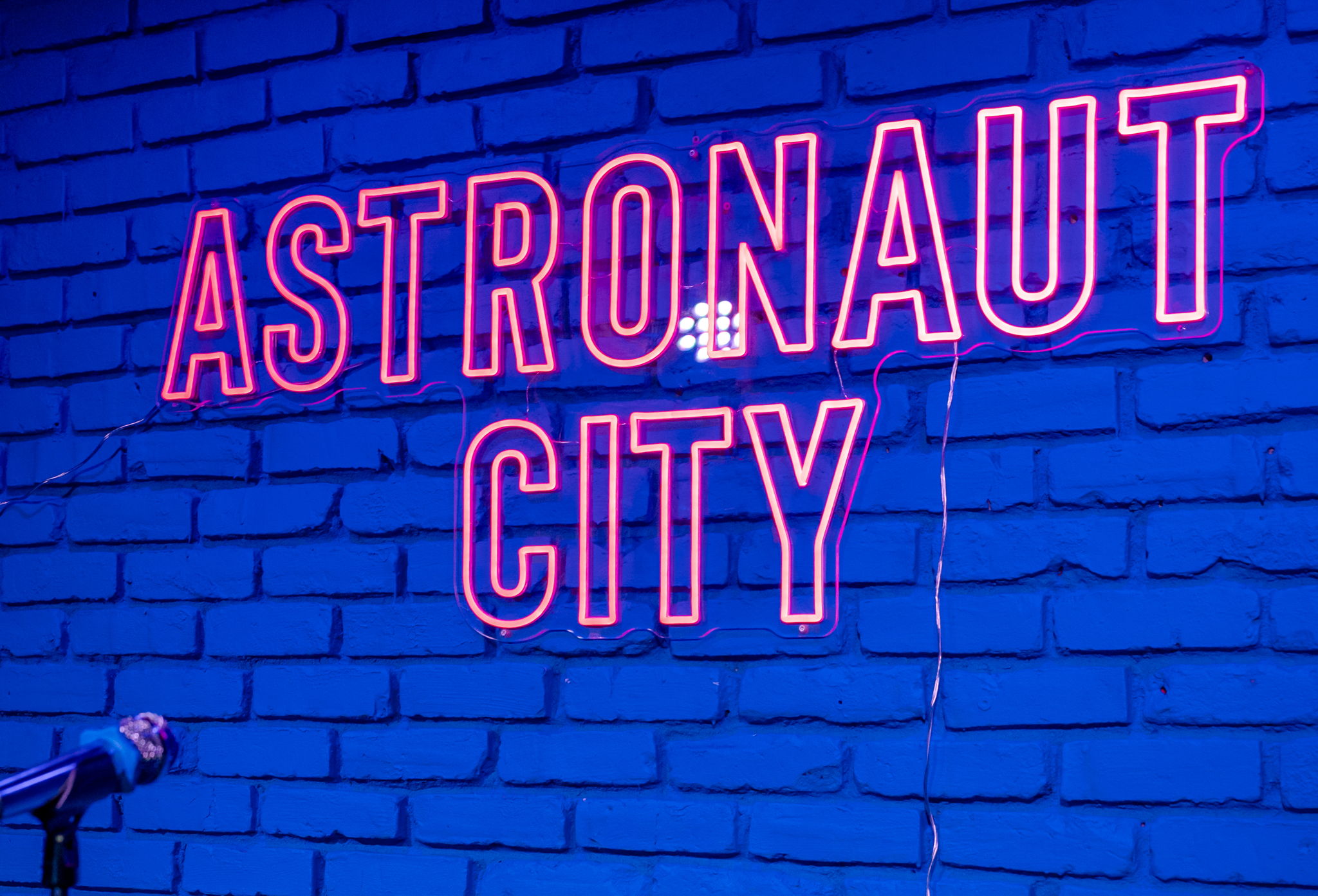 Neon sign reads 'ASTRONAUT CITY' on a brick wall, with a small astronaut figure hanging from the letter 'R'.