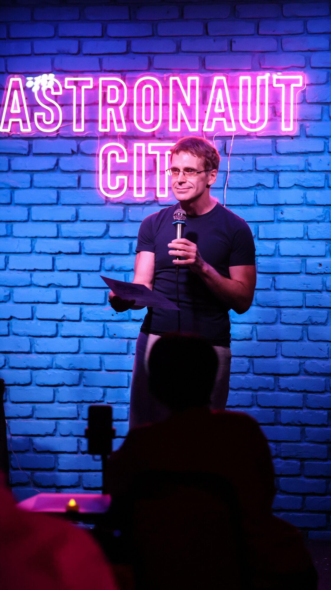 Person standing on stage holding a microphone and a tablet, in front of a blue brick wall with neon sign reading 'ASTRONAUT CITY'.