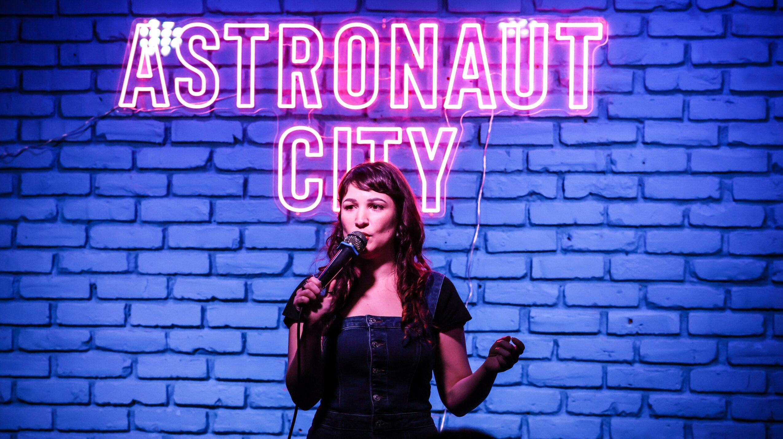 Woman singing into microphone in front of a blue brick wall with neon sign reading 'ASTRONAUT CITY'.