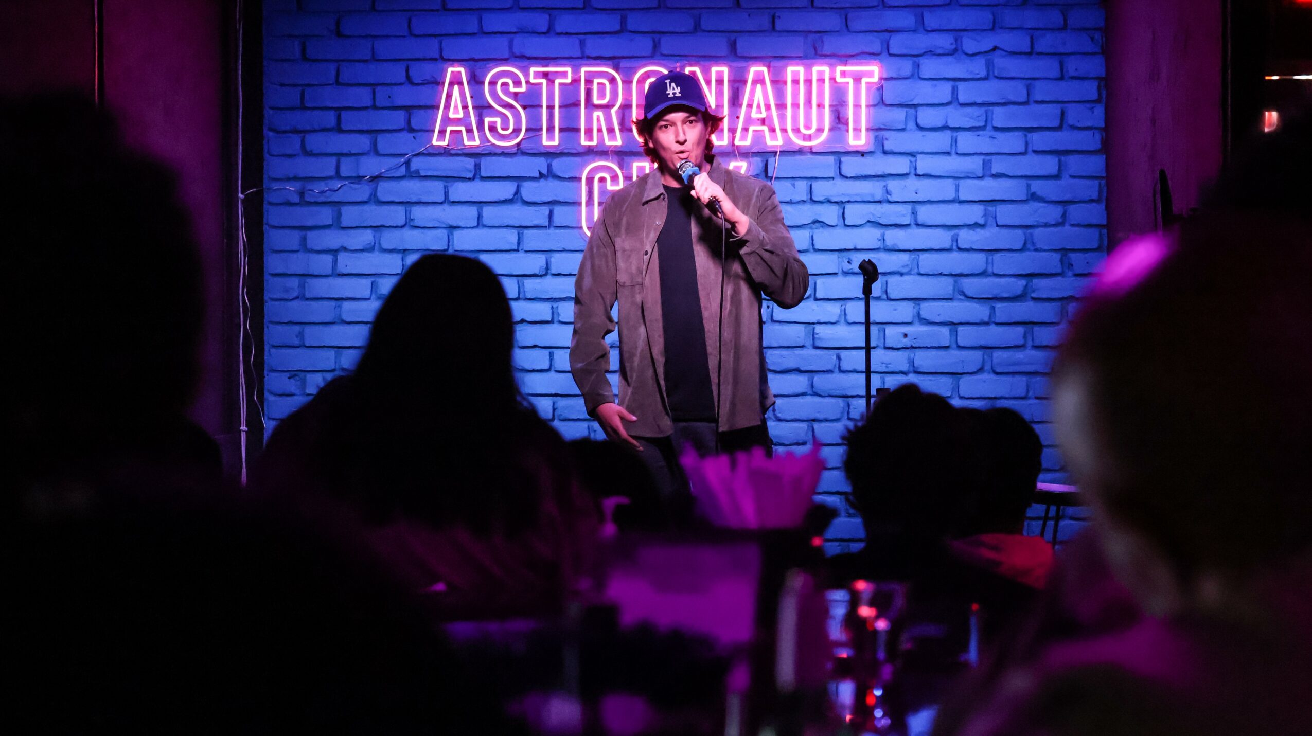 Person performing stand-up comedy on stage with a blue brick wall and neon sign reading 'ASTRONAUT' behind.