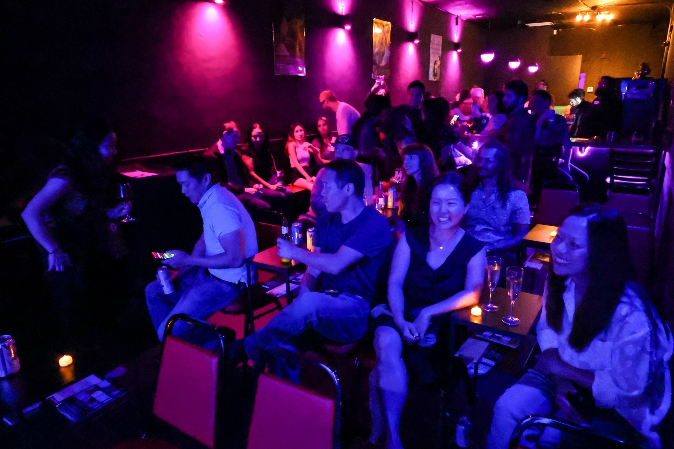 People sitting at tables in a dimly lit bar or club with purple and pink lighting, socializing and drinking.