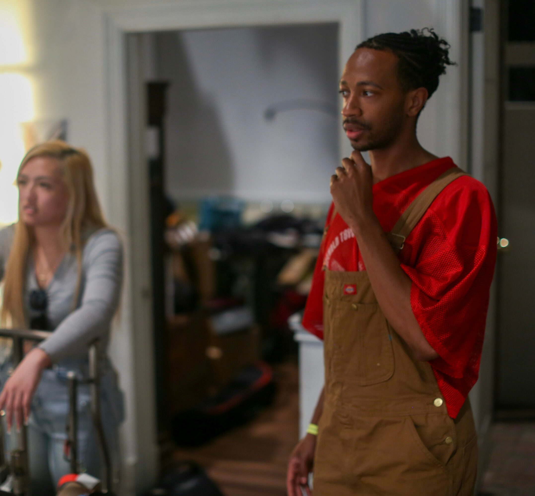 Man with dreadlocks wearing red shirt and brown overalls, standing indoors with hand on chin, looking thoughtful.