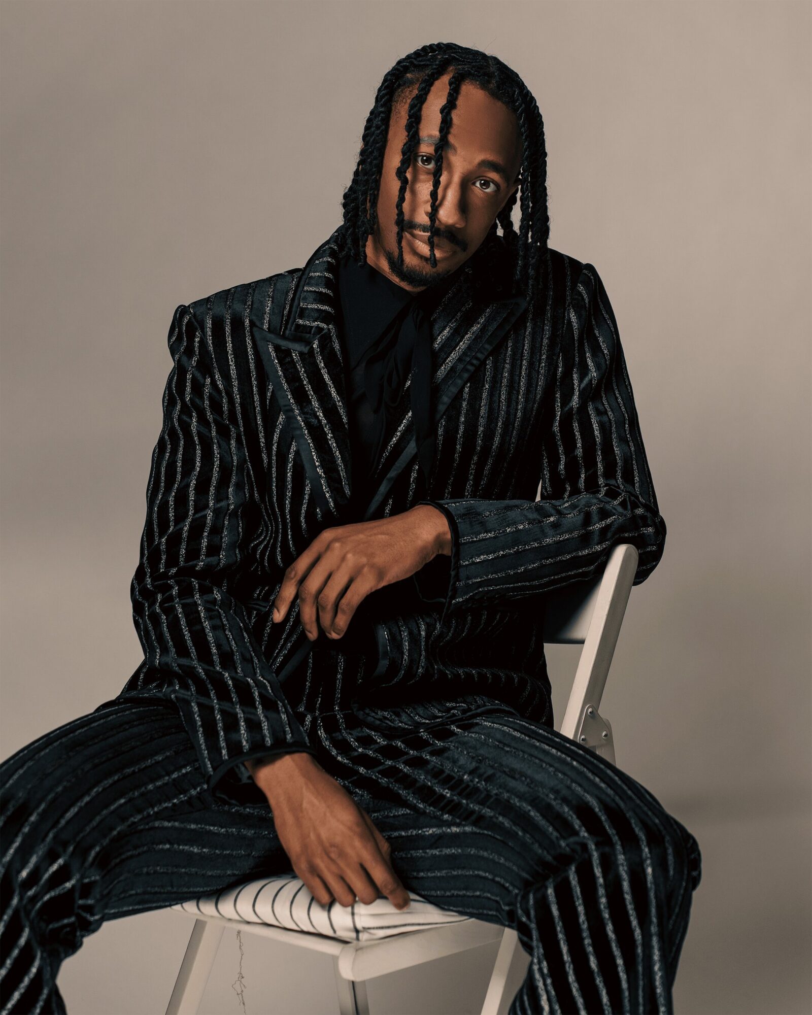 Person with braided hair wearing a pinstripe suit, sitting on a chair against a plain background.