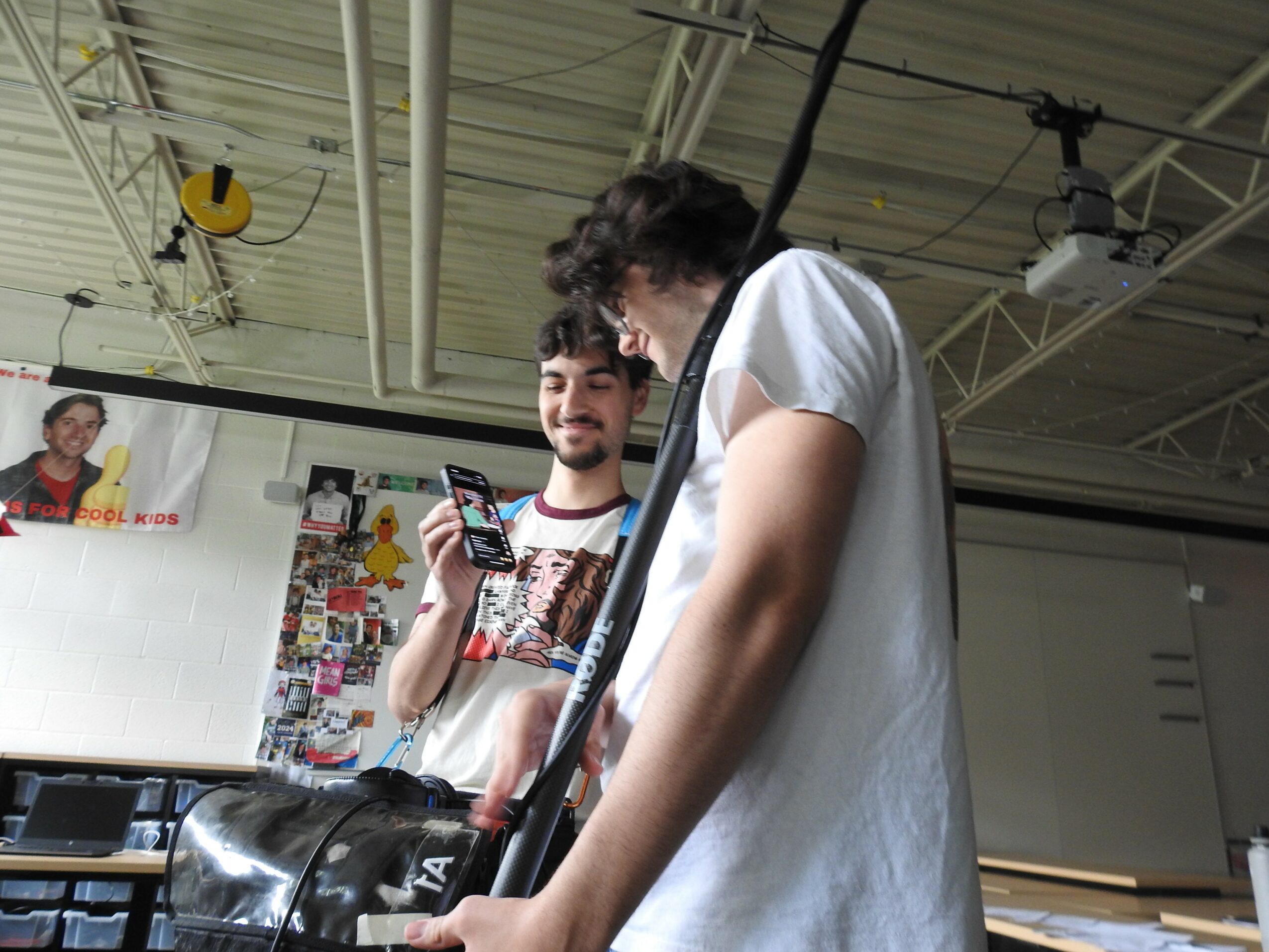 Two young men in a room with a high ceiling, one looking at a phone, the other with a bag, smiling.
