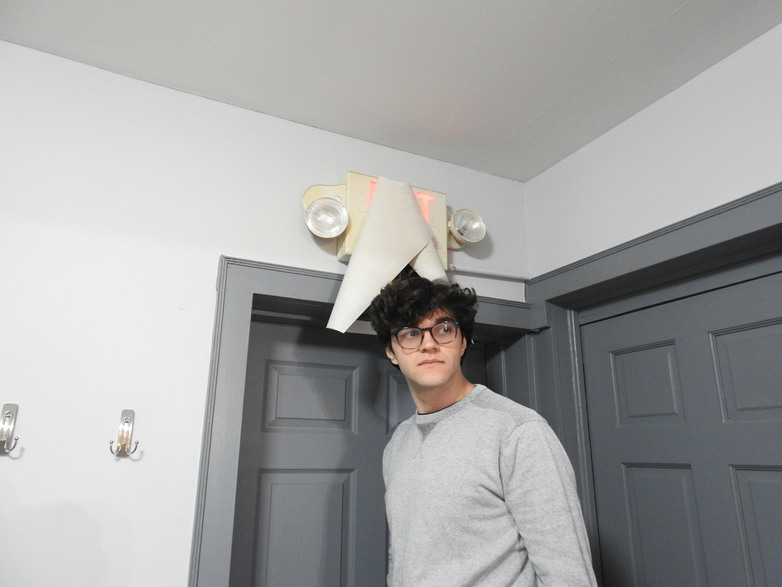 Young person with dark curly hair and glasses standing indoors near gray doors, wearing a light gray sweater.