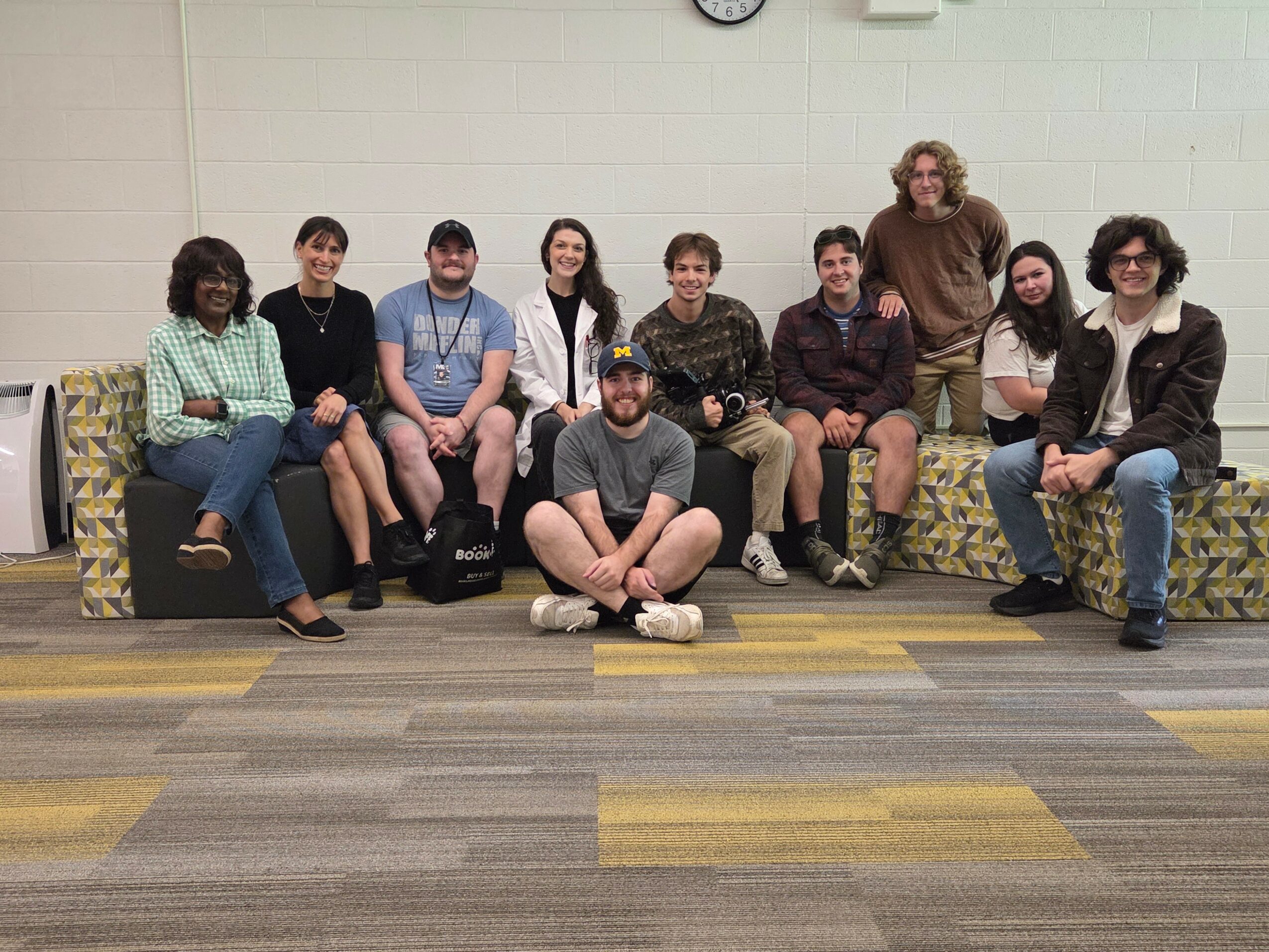 Group of ten people sitting and standing on a couch in a room with a white wall and a clock.