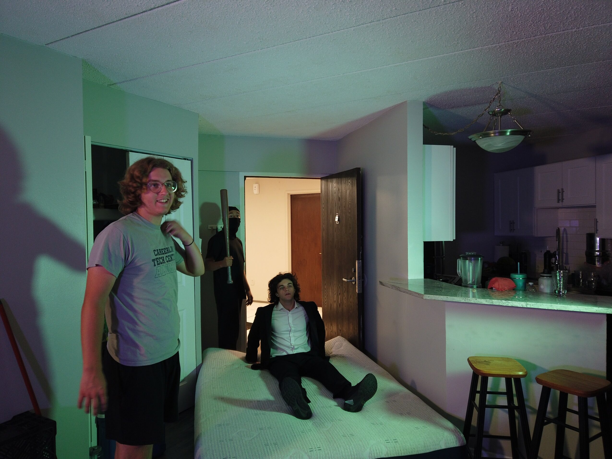 Person with curly hair and glasses standing near a bed in a room with green walls, a door, and a kitchen area.